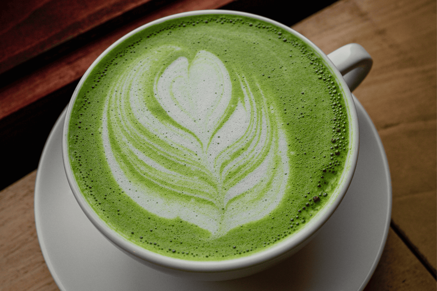 Matcha latte in a white cup and saucer with leaf-patterned latte art, on a wooden table
