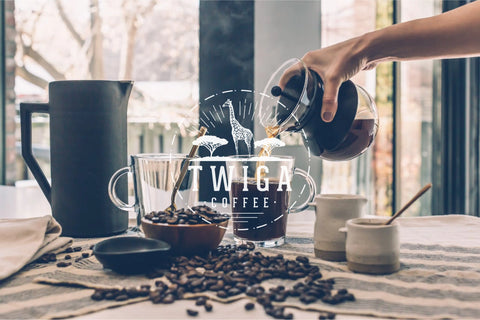Freshly brewed coffee being poured from a carafe into a glass mug, with beans and a French press on the table.
